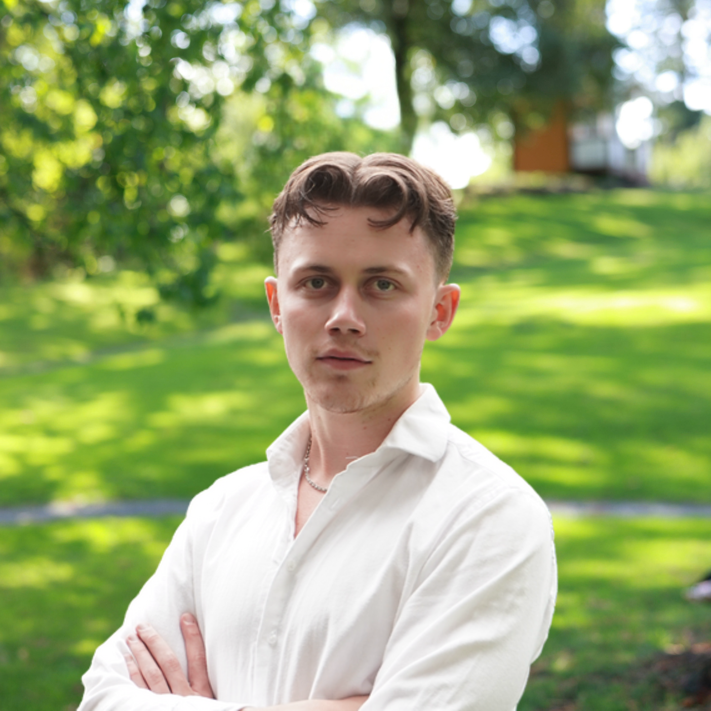 A young man with short brown hair wearing a white button-down shirt stands with his arms crossed in a park.