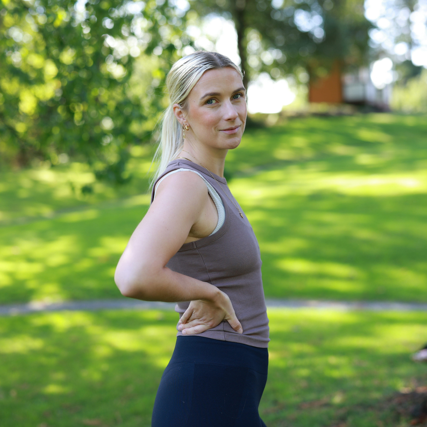 A woman with blonde hair in a ponytail wearing athletic wear stands in a park and looks toward the camera.