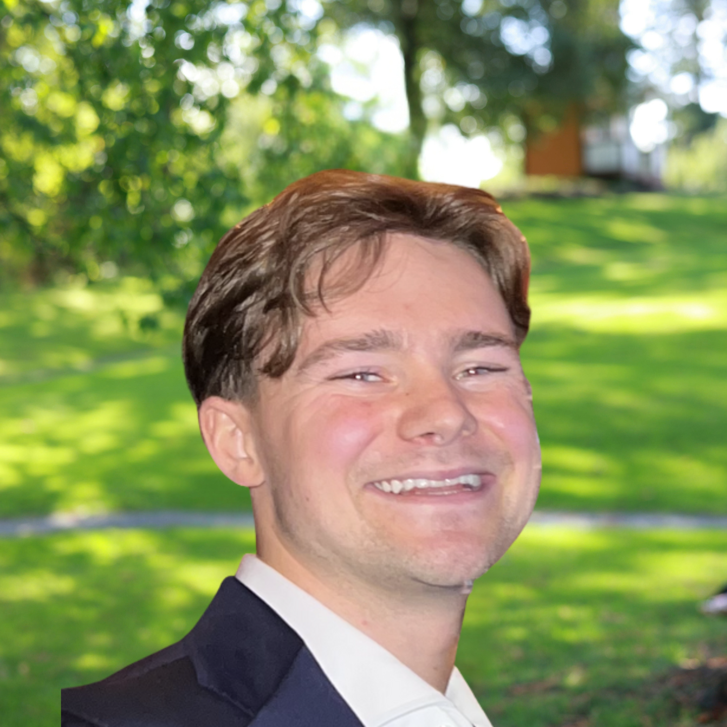 A young man with brown hair smiles warmly while wearing a suit jacket in a park setting.