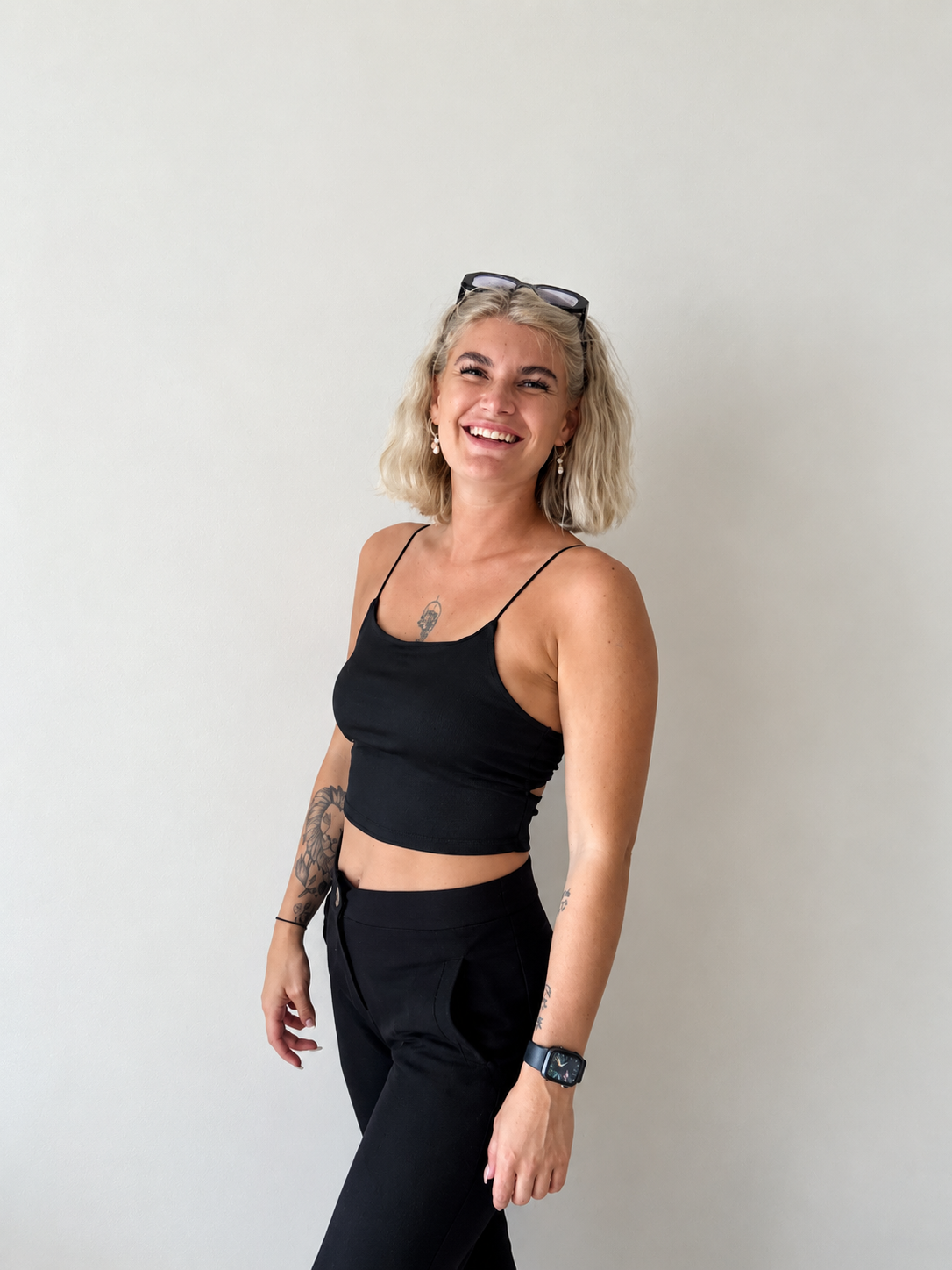 A smiling blonde woman with tattoos wearing a black crop top and pants stands against a plain white wall.