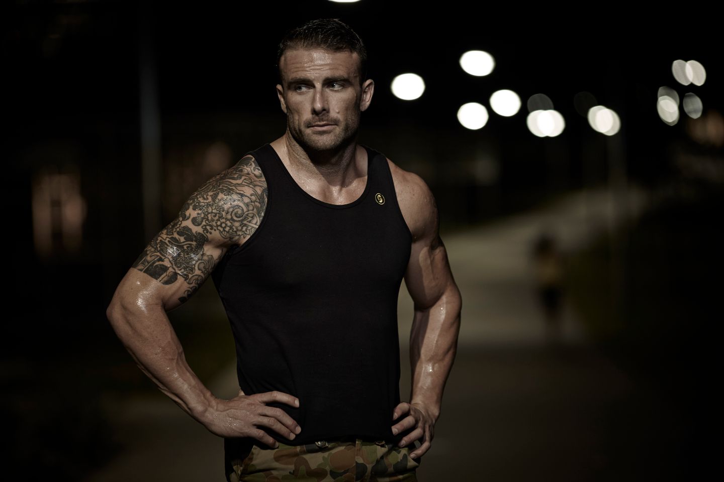 A muscular man with a tattooed arm stands with hands on his hips outdoors at night with blurred city lights behind him.