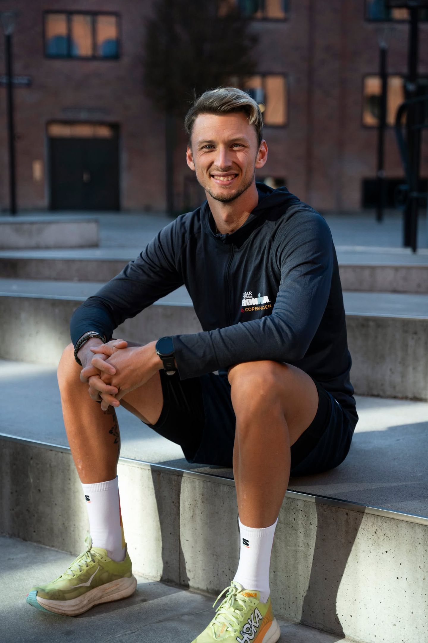 A smiling man in athletic gear sits on concrete steps outdoors.