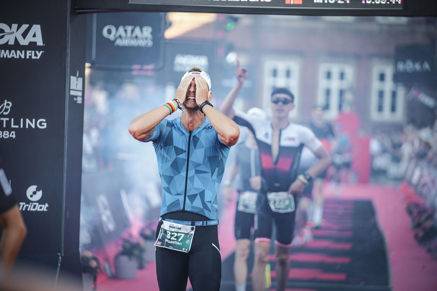 A triathlete crosses the finish line with his hands over his face in an emotional display of exhaustion and triumph.