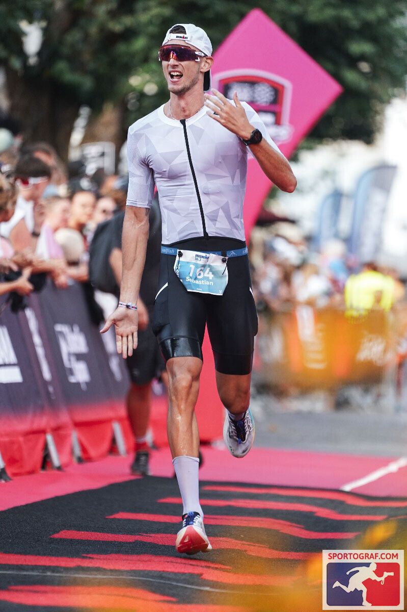 A triathlete in a white jersey and black shorts celebrates while running across the finish line of a race.