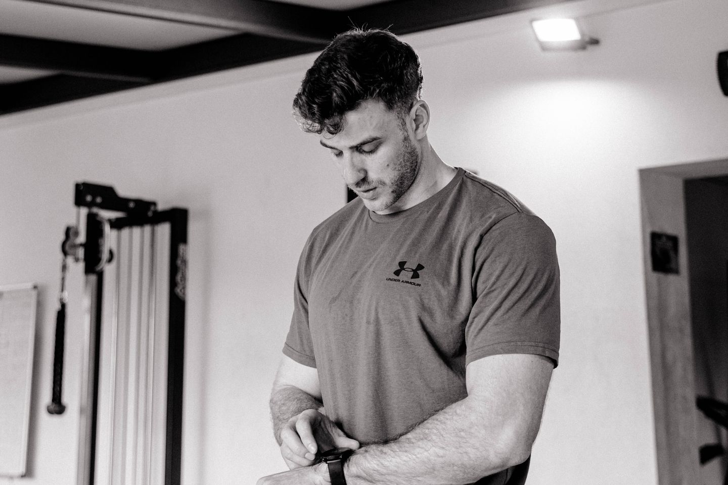 A man in a t-shirt checks his fitness tracker while standing in a gym.