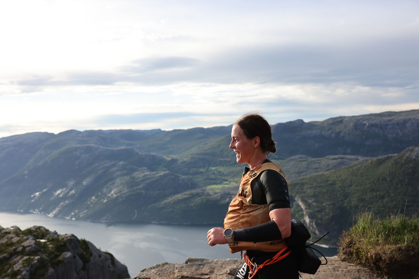 A woman wearing a hydration vest trail runs along a mountain ridge overlooking a large fjord.
