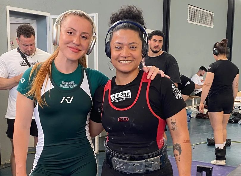 Two female powerlifters wearing headphones and athletic gear pose together for a photo in a gym.