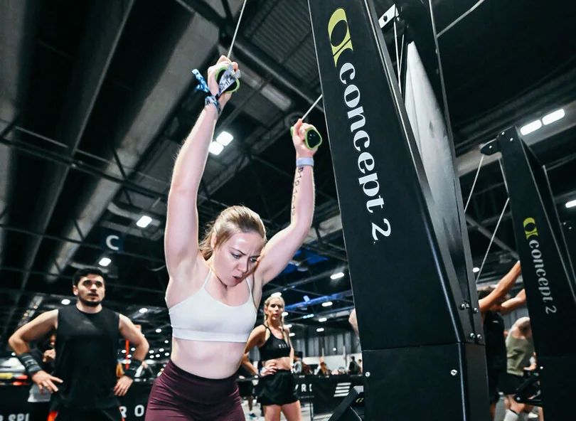 A woman in a gym focuses intensely while using a ski erg machine with her arms raised.