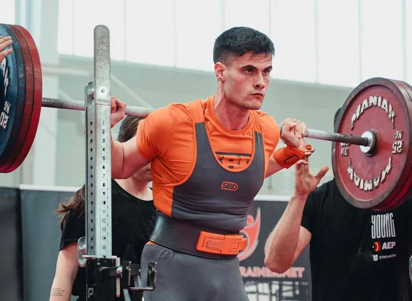 A focused athlete wearing a lifting singlet and belt prepares to perform a heavy barbell squat in a gym.