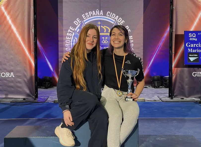 Two young women smiling together on a competition podium, with one holding a trophy and wearing a medal.