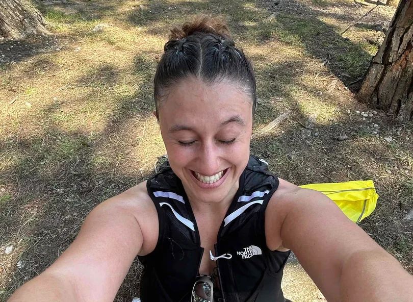 A smiling woman with braided hair wearing a running vest takes a selfie while outdoors in a wooded area.
