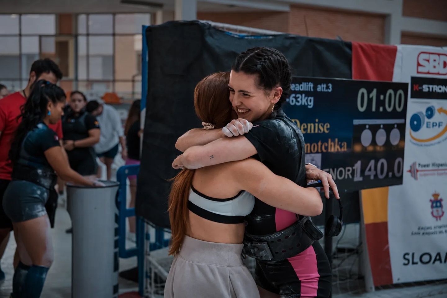 Two women embrace in celebration at a powerlifting competition with a scoreboard visible in the background.