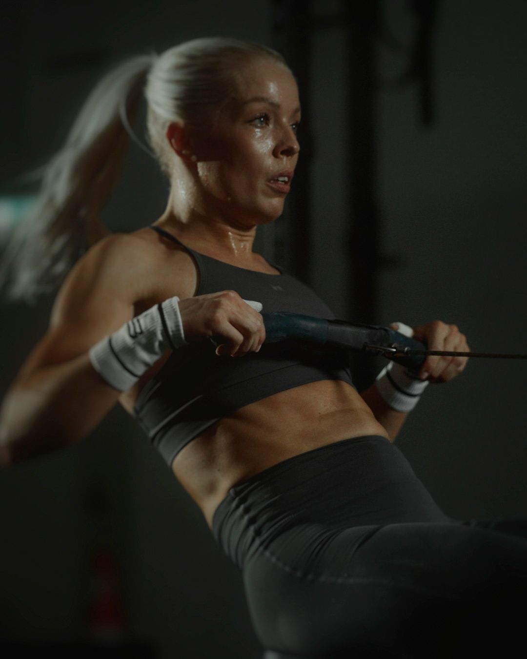 A focused woman with a ponytail performs a rowing exercise in a dimly lit gym.