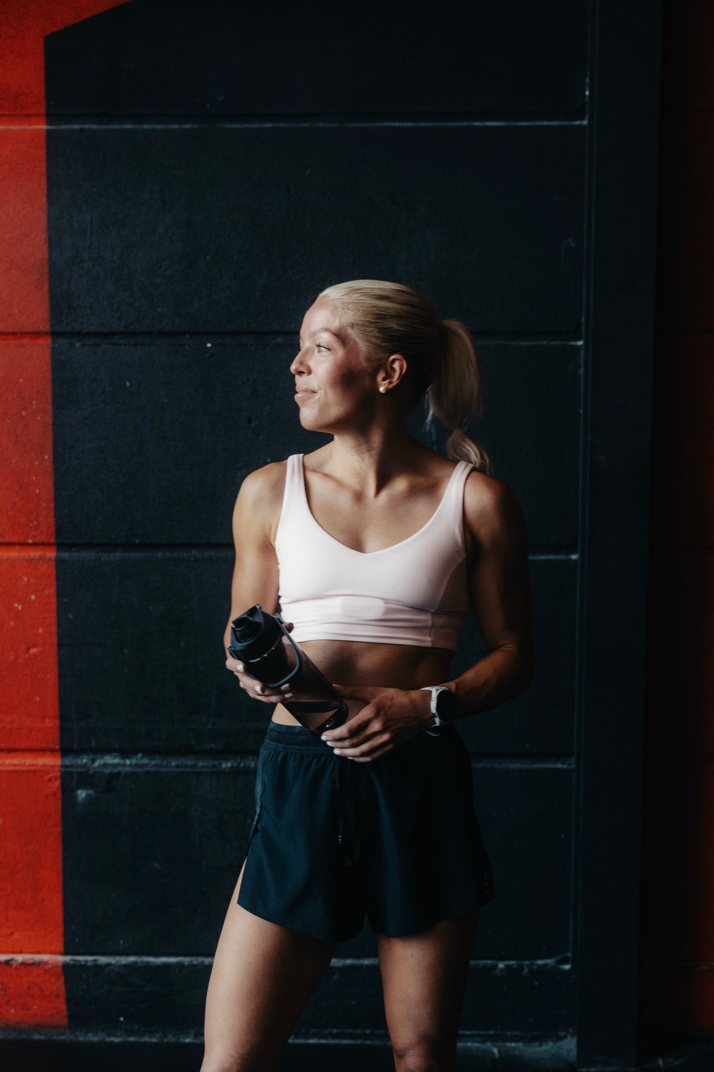 A blonde woman in athletic wear holds a water bottle while looking away in a gym.