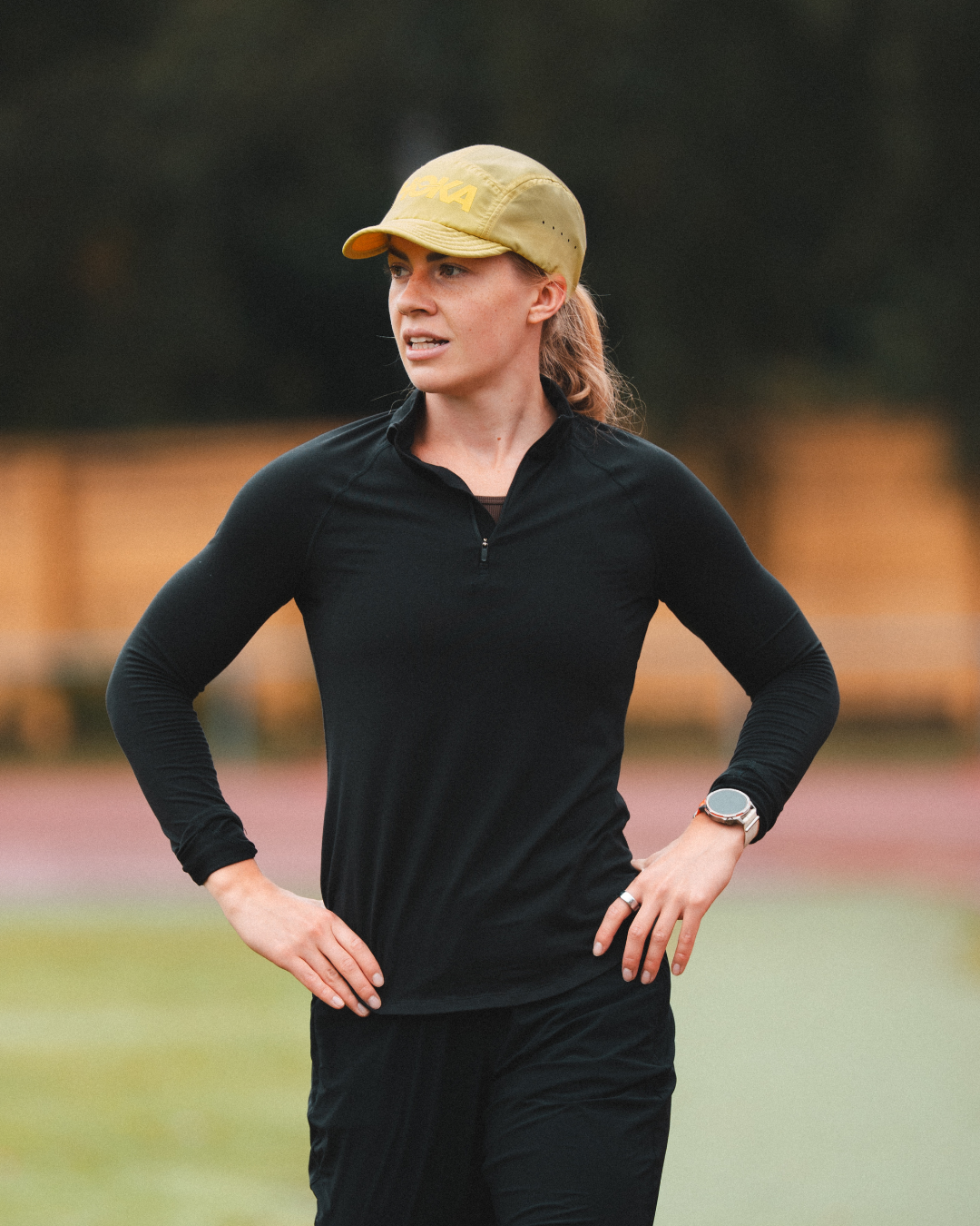 A woman in a black long-sleeve shirt and yellow cap stands on a track with her hands on her hips.