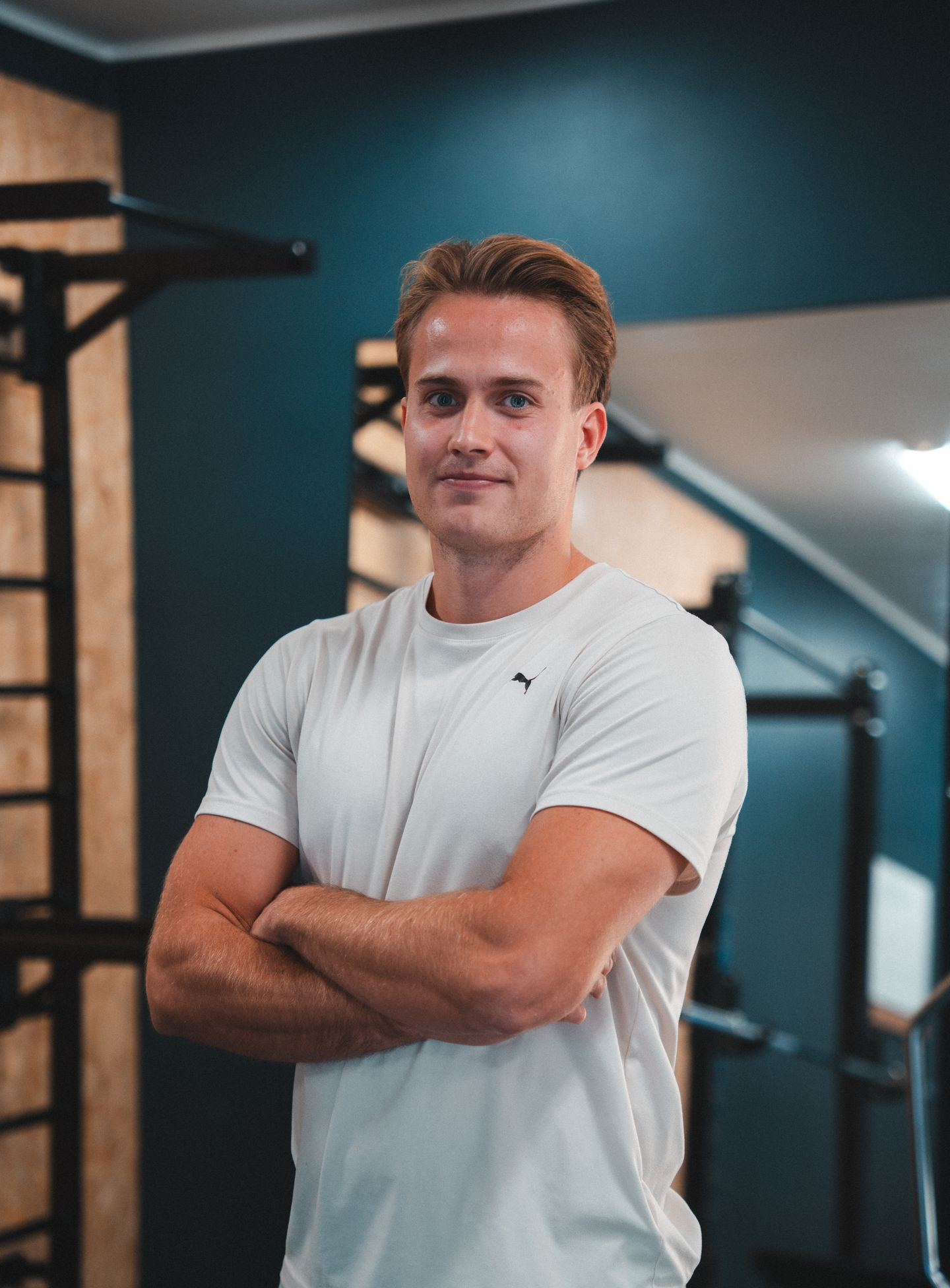 A fit man in a white t-shirt stands with his arms crossed in a gym.