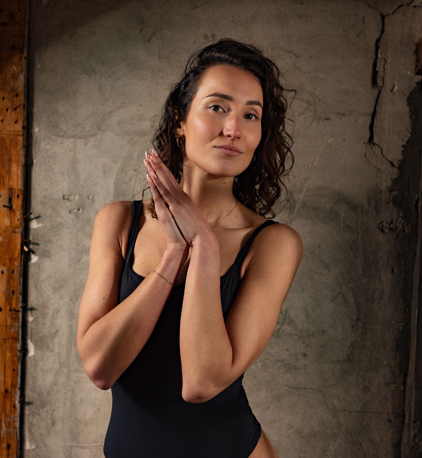 A woman with curly hair wearing a black bodysuit poses with her hands pressed together against a textured concrete wall.