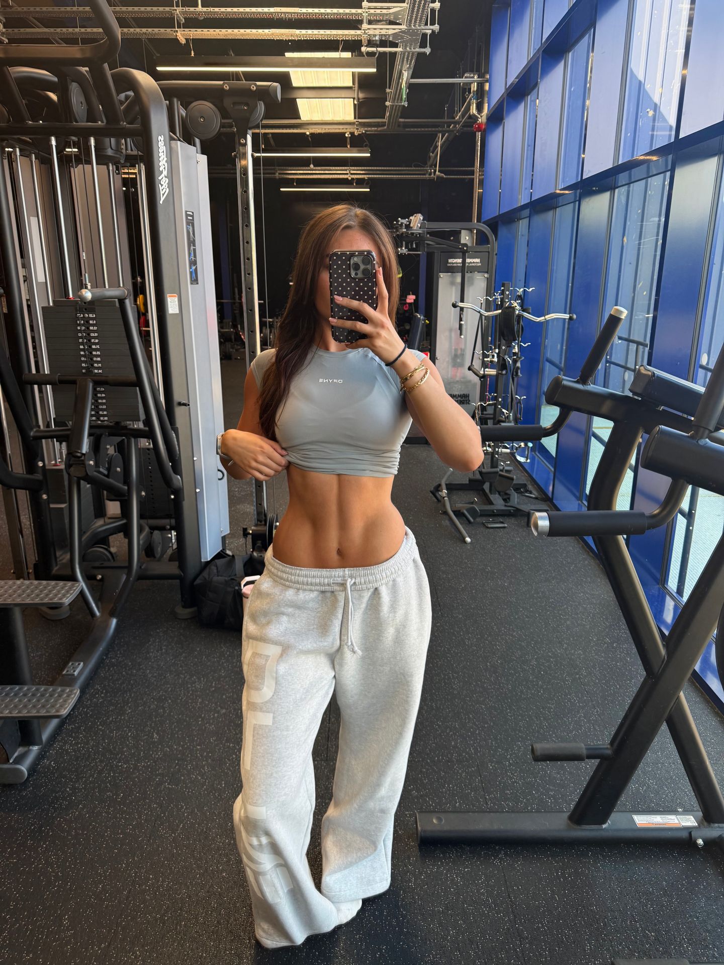 A woman in a cropped top and sweatpants takes a mirror selfie in a gym.