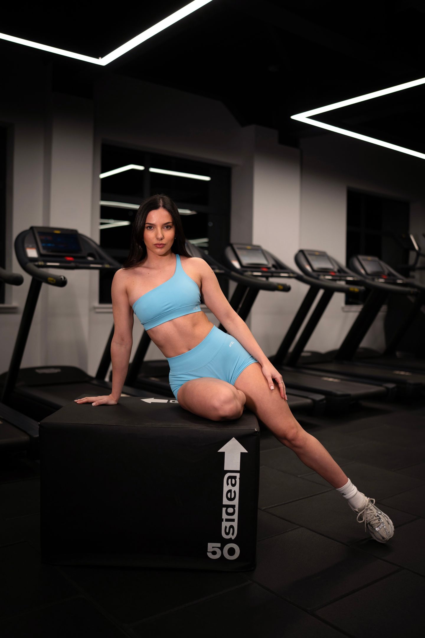 A woman in a light blue athletic set sits on a black gym box in front of a row of treadmills.