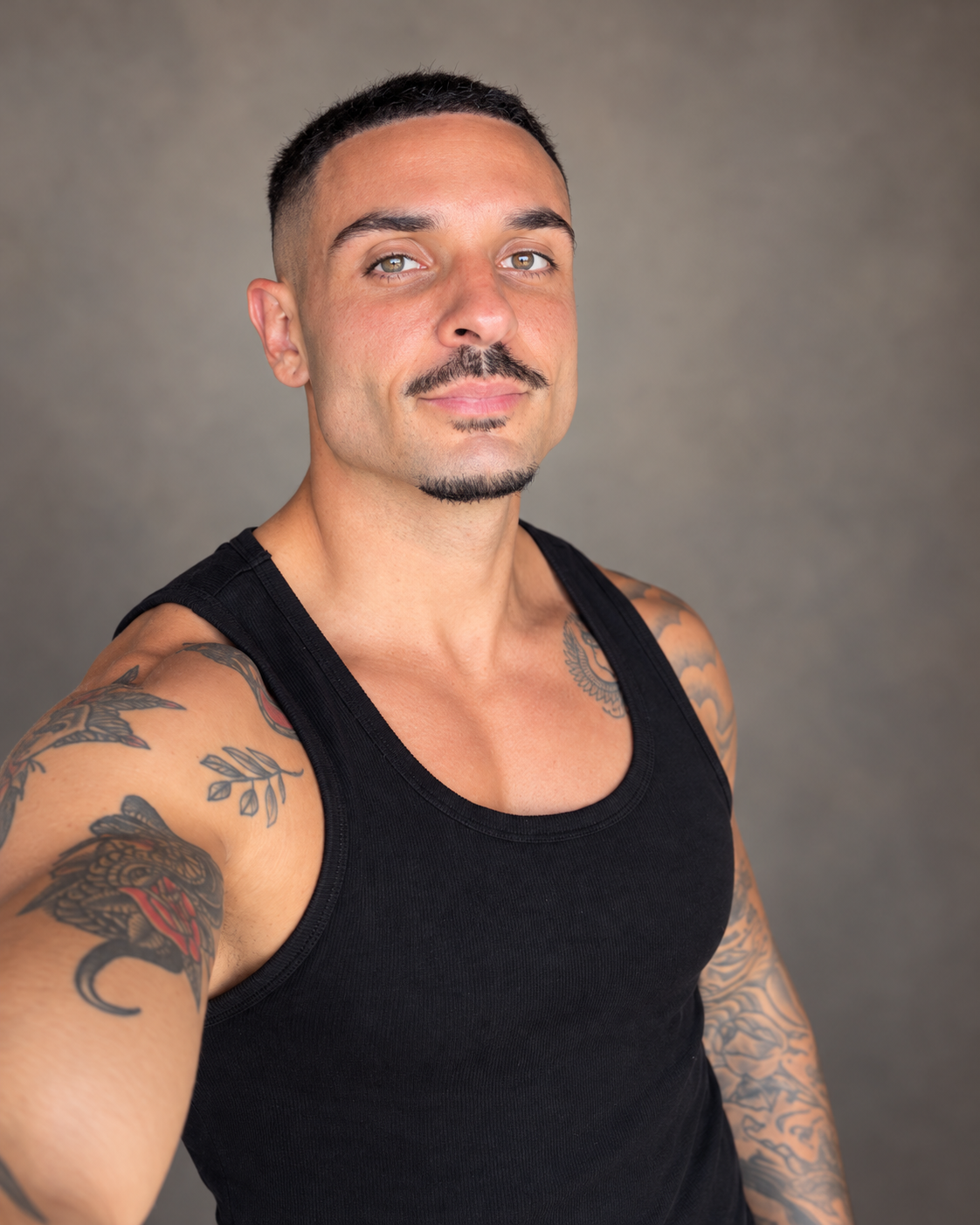 A tattooed man with a short haircut and a black tank top poses against a neutral gray background.