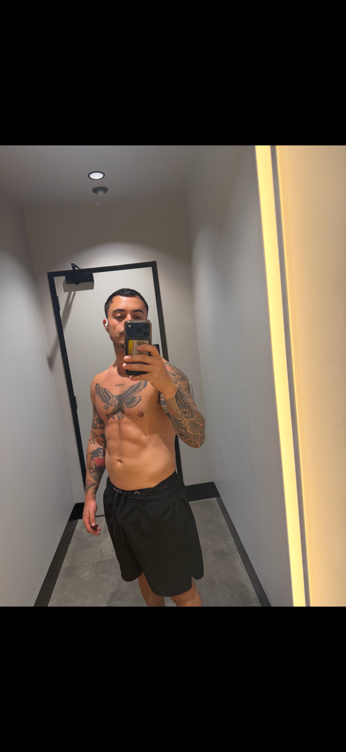 A shirtless man with tattoos on his chest and arms takes a mirror selfie in a gym locker room.