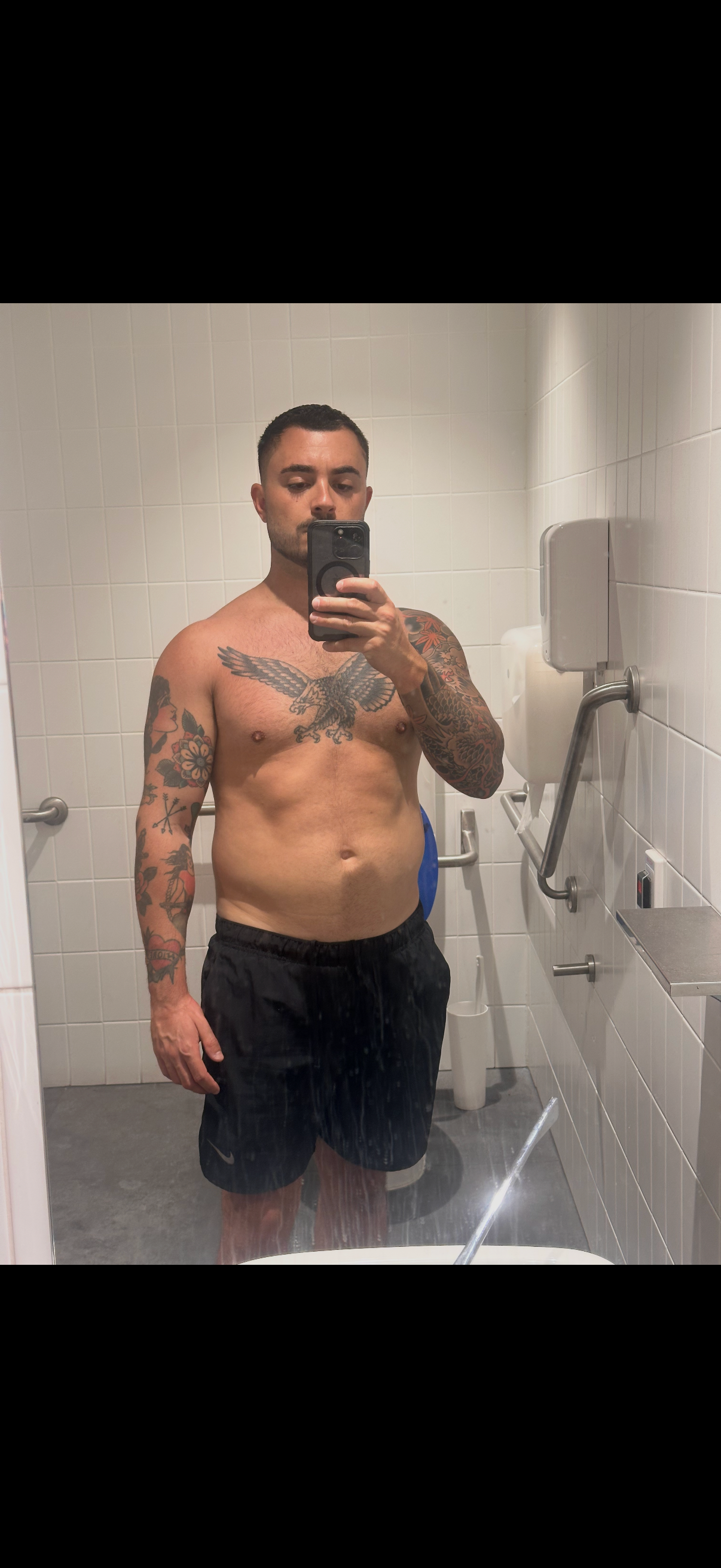 A shirtless man with tattoos on his chest and arms takes a mirror selfie in a tiled bathroom.