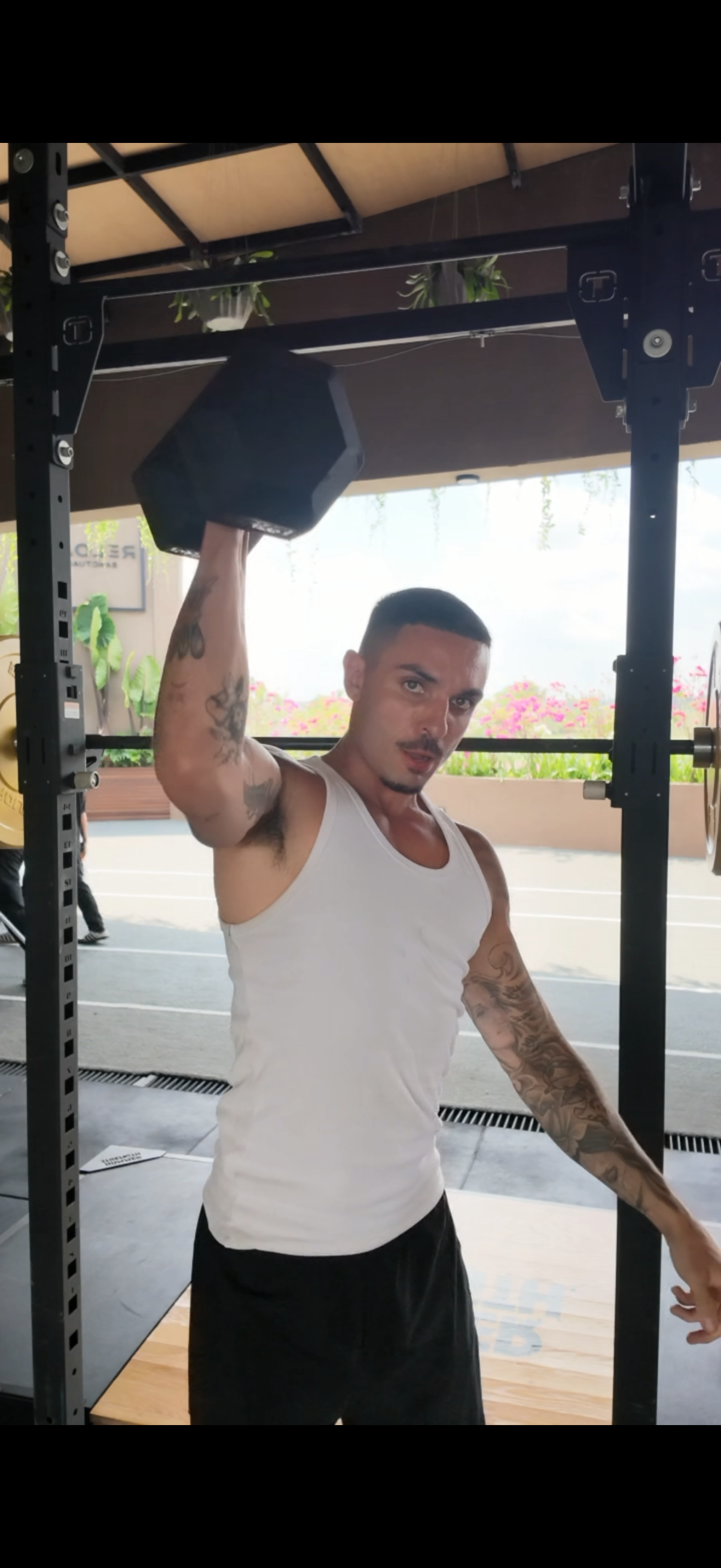 A man in a white tank top performs a single-arm dumbbell overhead press in an open-air gym.