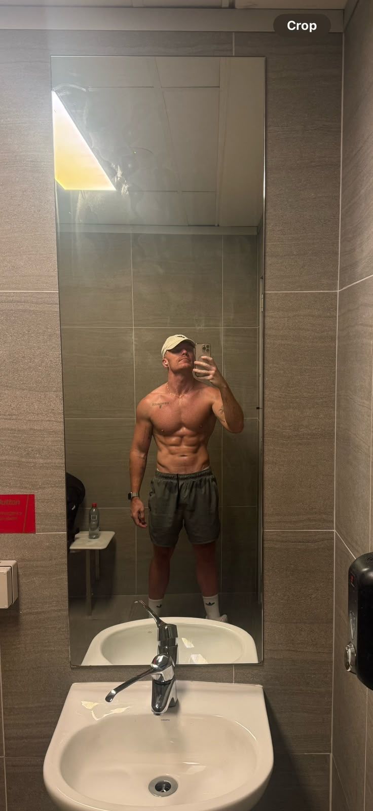 A shirtless man wearing a baseball cap and athletic shorts takes a mirror selfie in a gym bathroom.