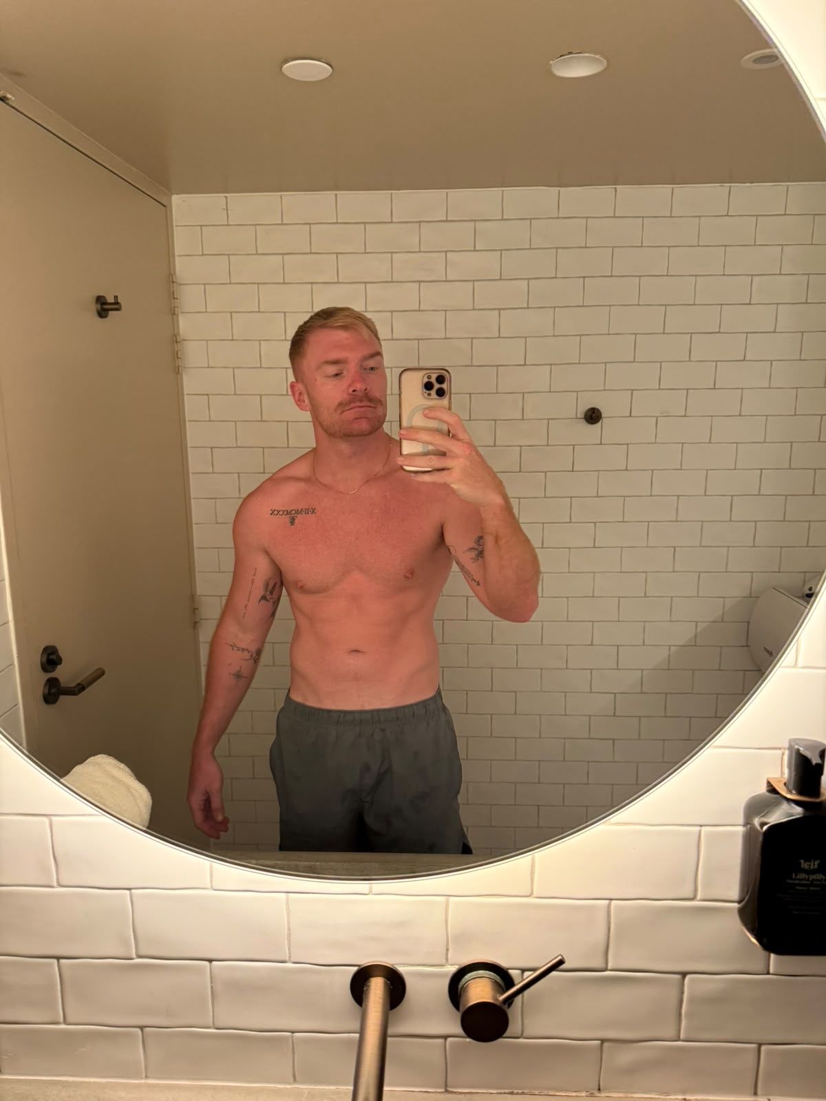 A shirtless man with tattoos takes a mirror selfie in a bathroom with white subway tile walls.