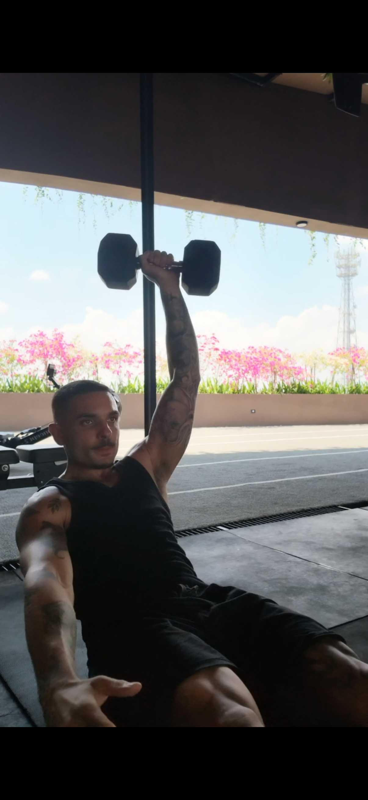A man in a black tank top performs a single-arm dumbbell overhead press while sitting in an open-air gym.