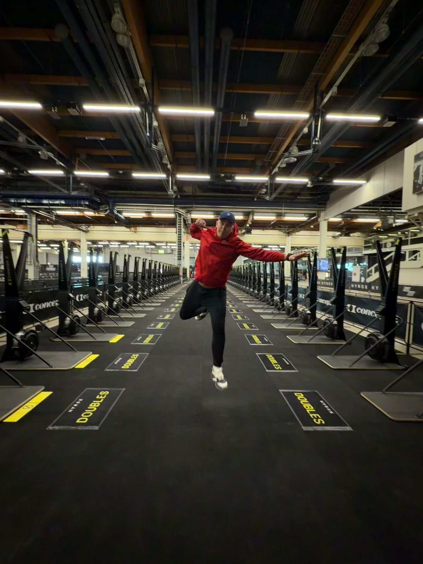 A man in a red hoodie jumps in the air in a large gym filled with rows of rowing machines.
