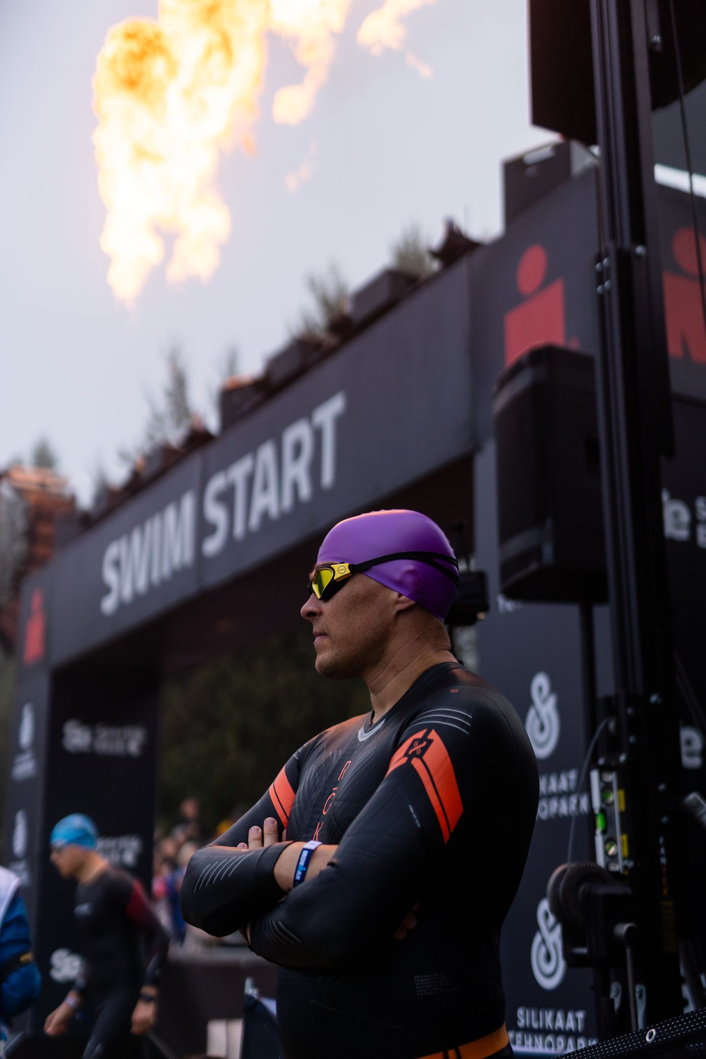 A triathlete in a wetsuit and purple swim cap stands with arms crossed at the swim start line with flames in the background.