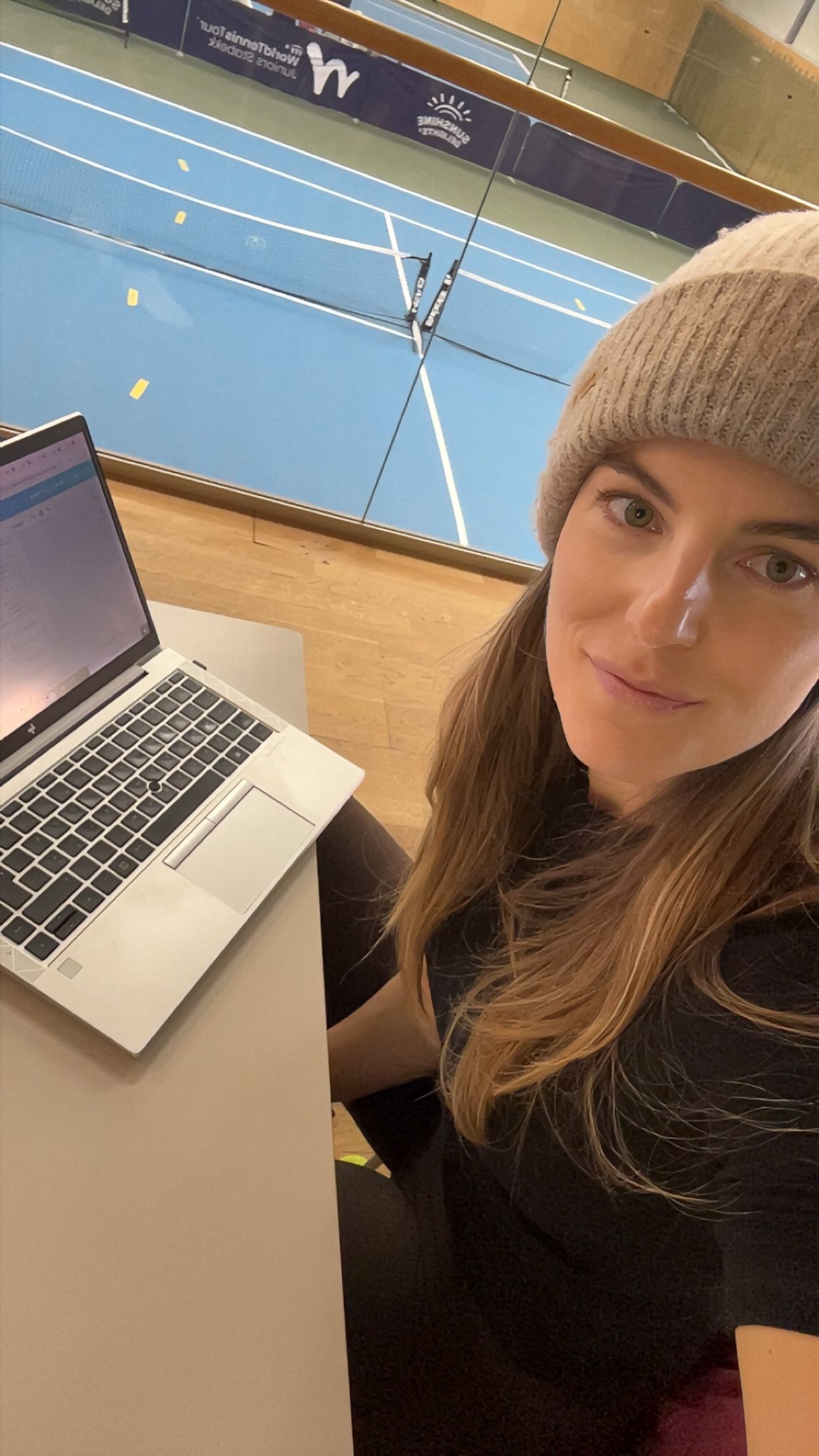 A woman wearing a beanie sits at a desk with a laptop, overlooking an indoor sports court.