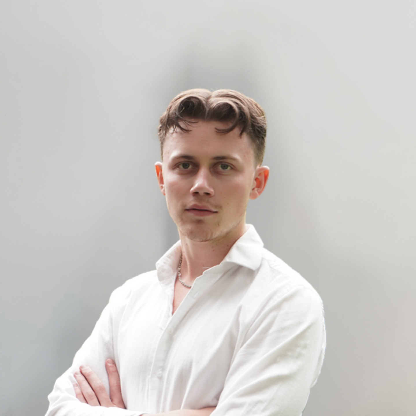 A young man with short brown hair wearing a white button-down shirt stands with his arms crossed against a plain background.