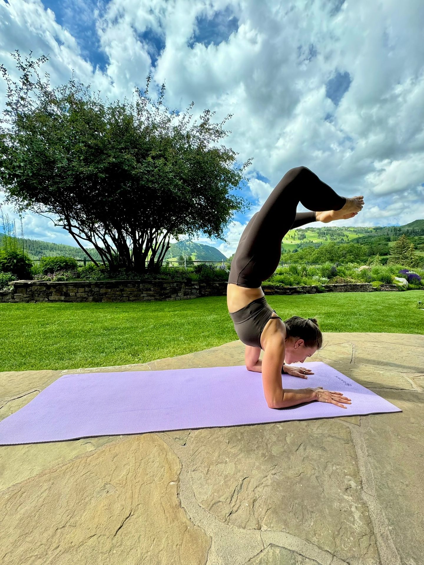 A woman performs a forearm stand with bent legs on a purple yoga mat in a lush outdoor garden setting.