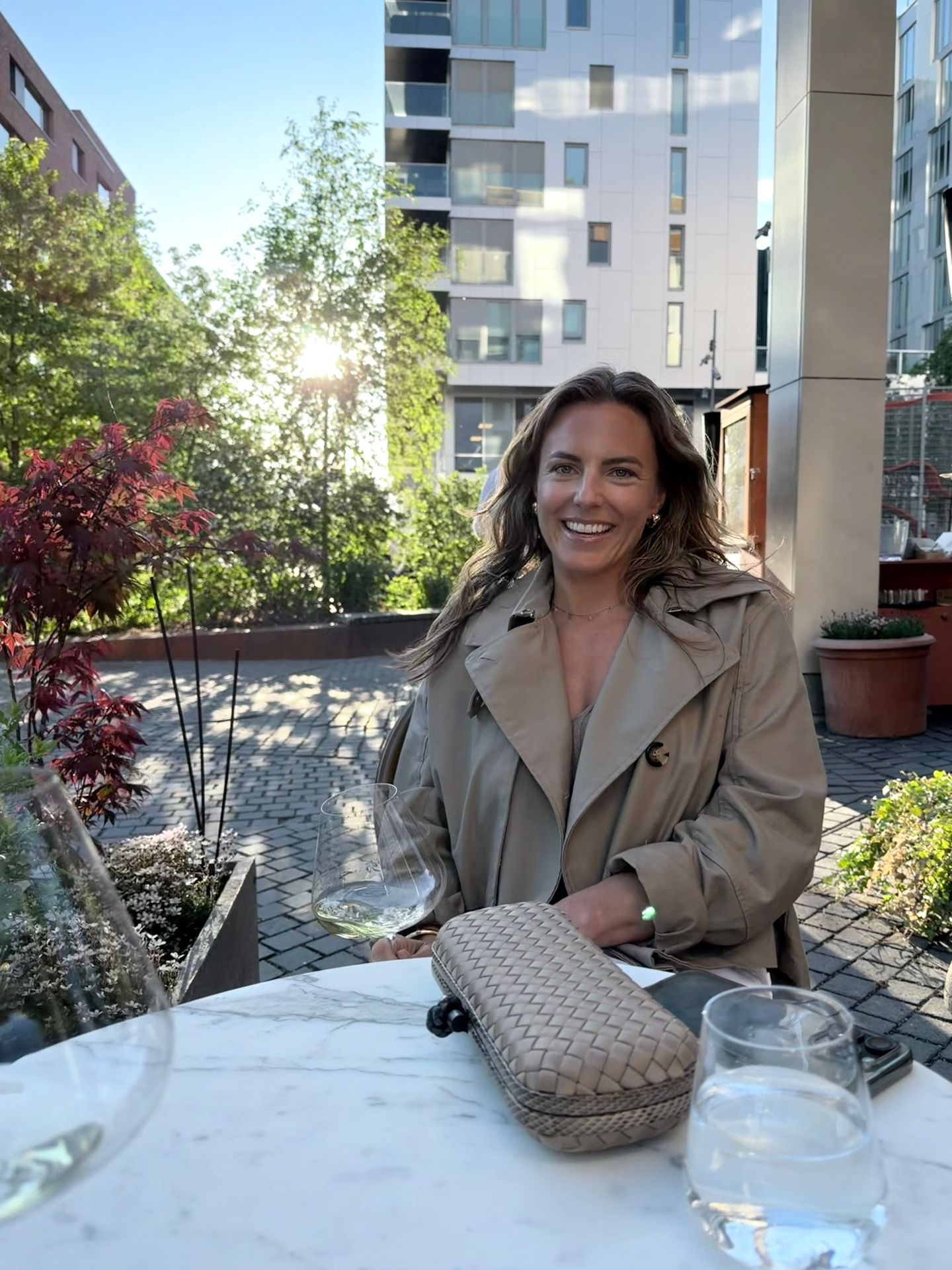 A smiling woman in a trench coat sits at an outdoor cafe table with a glass of wine.