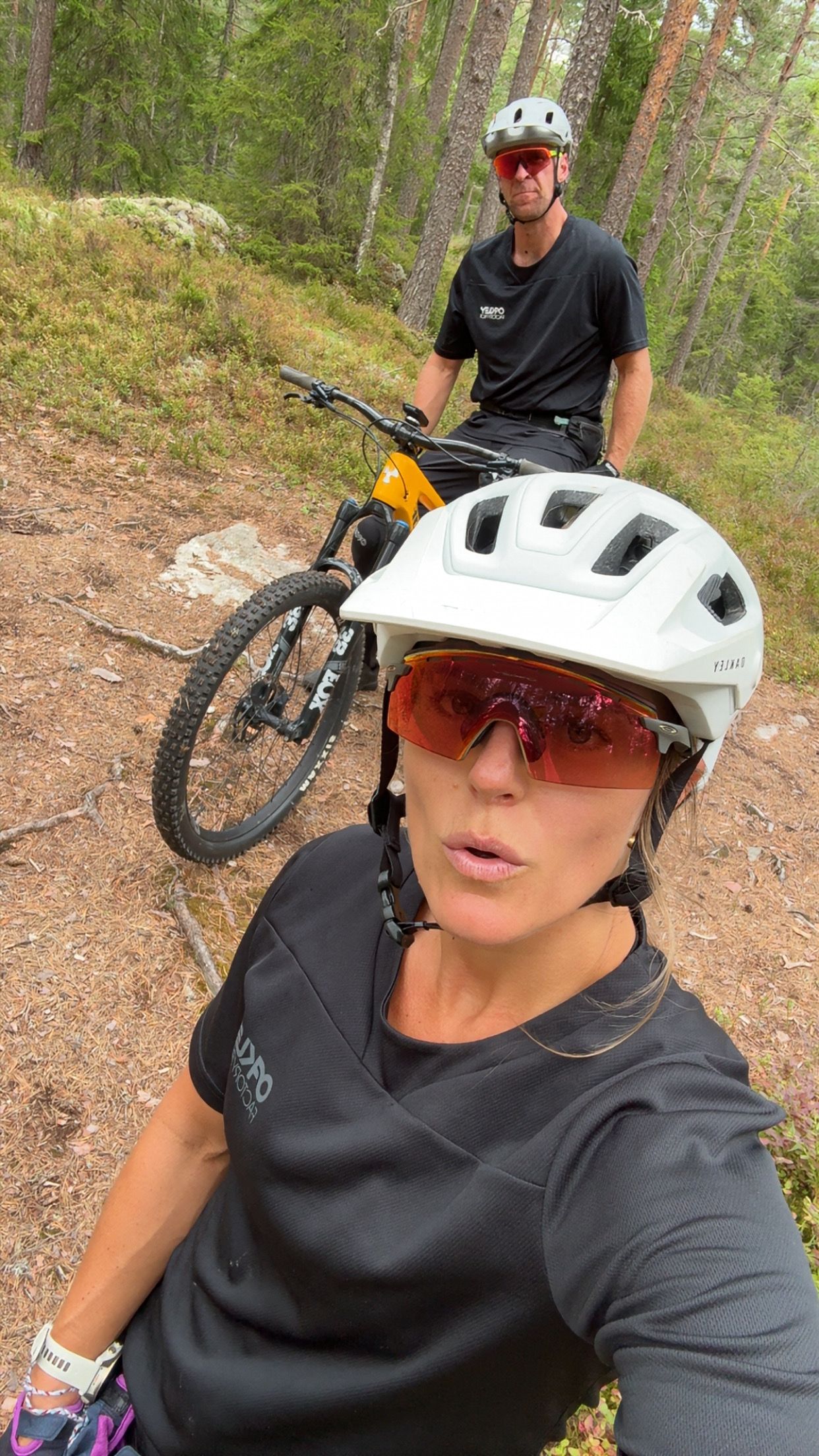 A woman and a man wearing cycling helmets and sunglasses take a selfie while mountain biking on a forest trail.