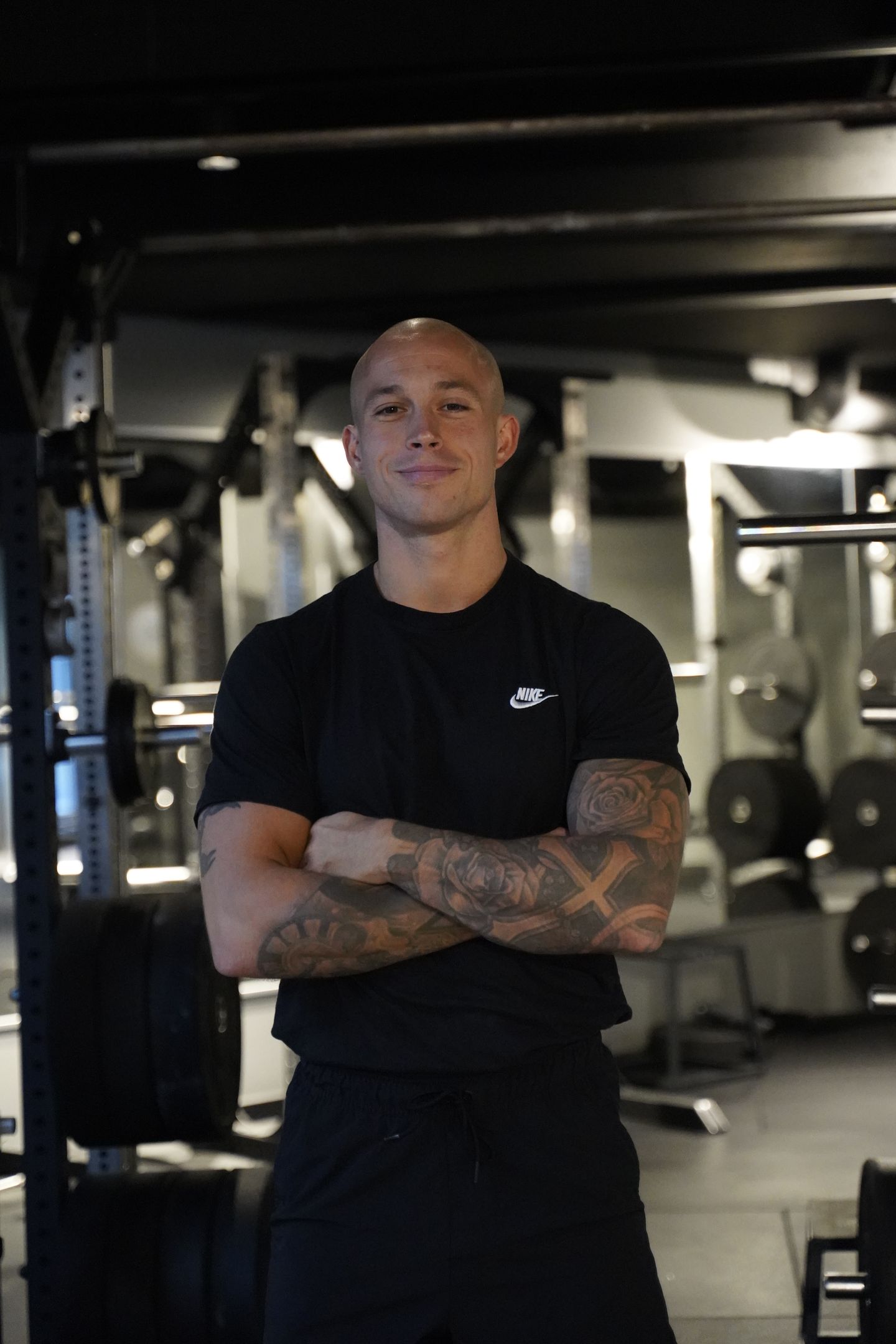 A bald man with tattooed arms stands with his arms crossed in a gym.