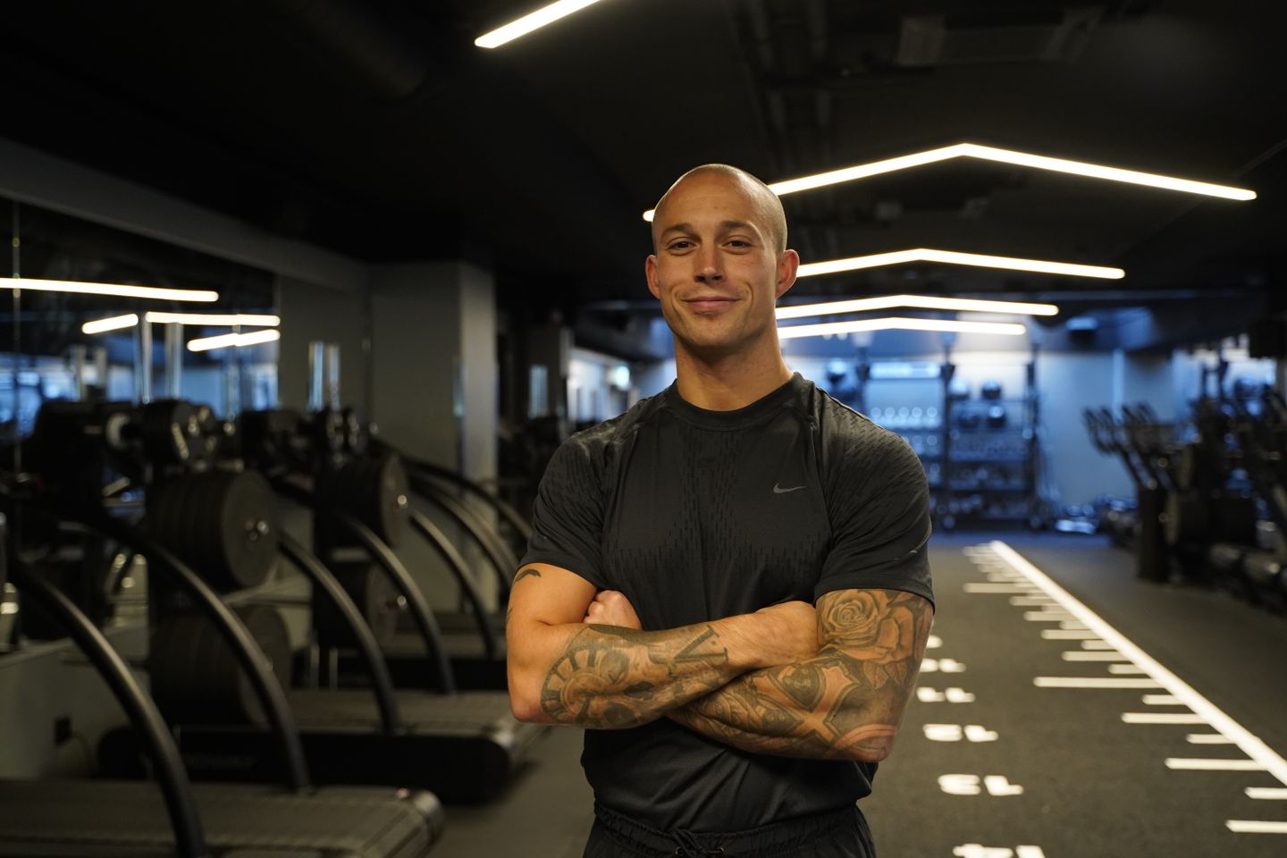 A bald, tattooed fitness coach stands with arms crossed in a modern gym.