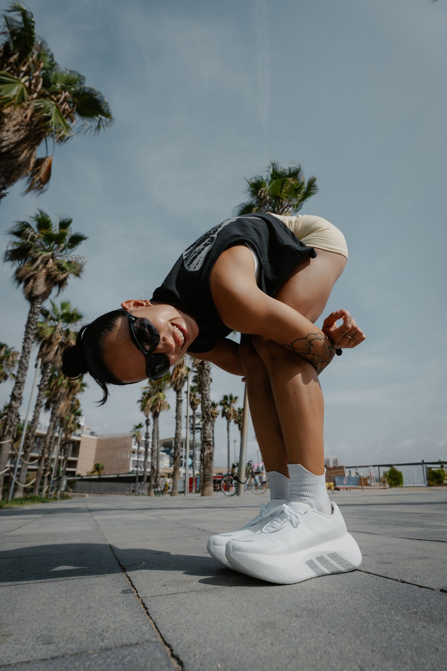 A woman in athletic wear bends forward at the waist while smiling on a paved path lined with palm trees.