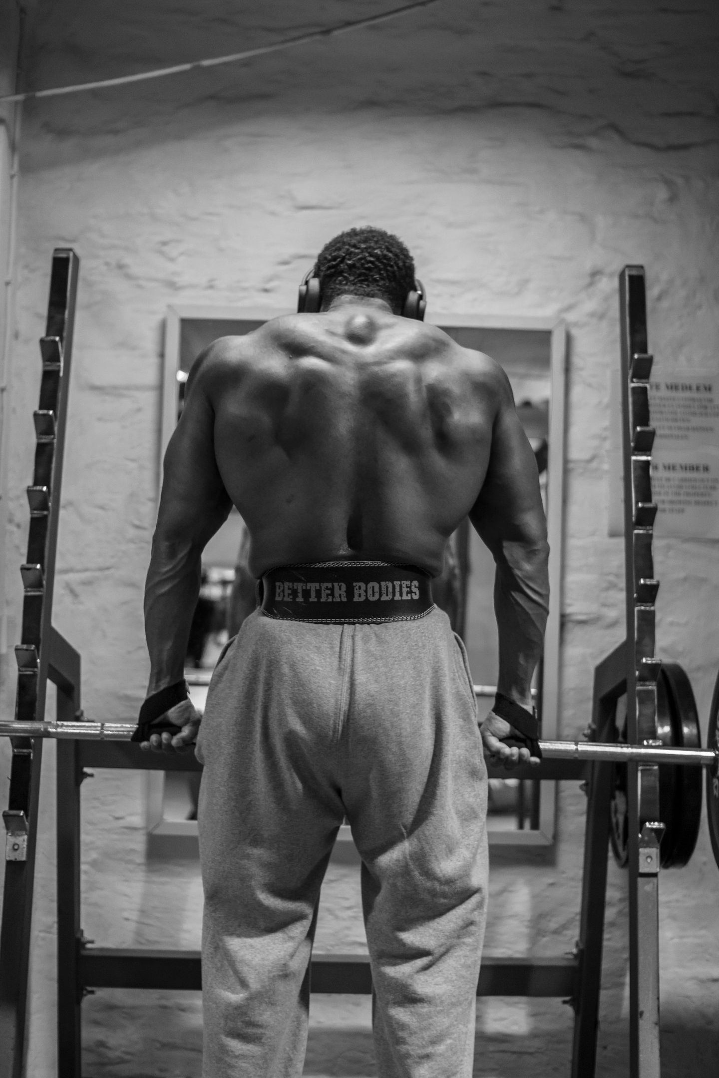 A muscular man with his back to the camera stands in a gym holding a barbell while wearing a weightlifting belt.