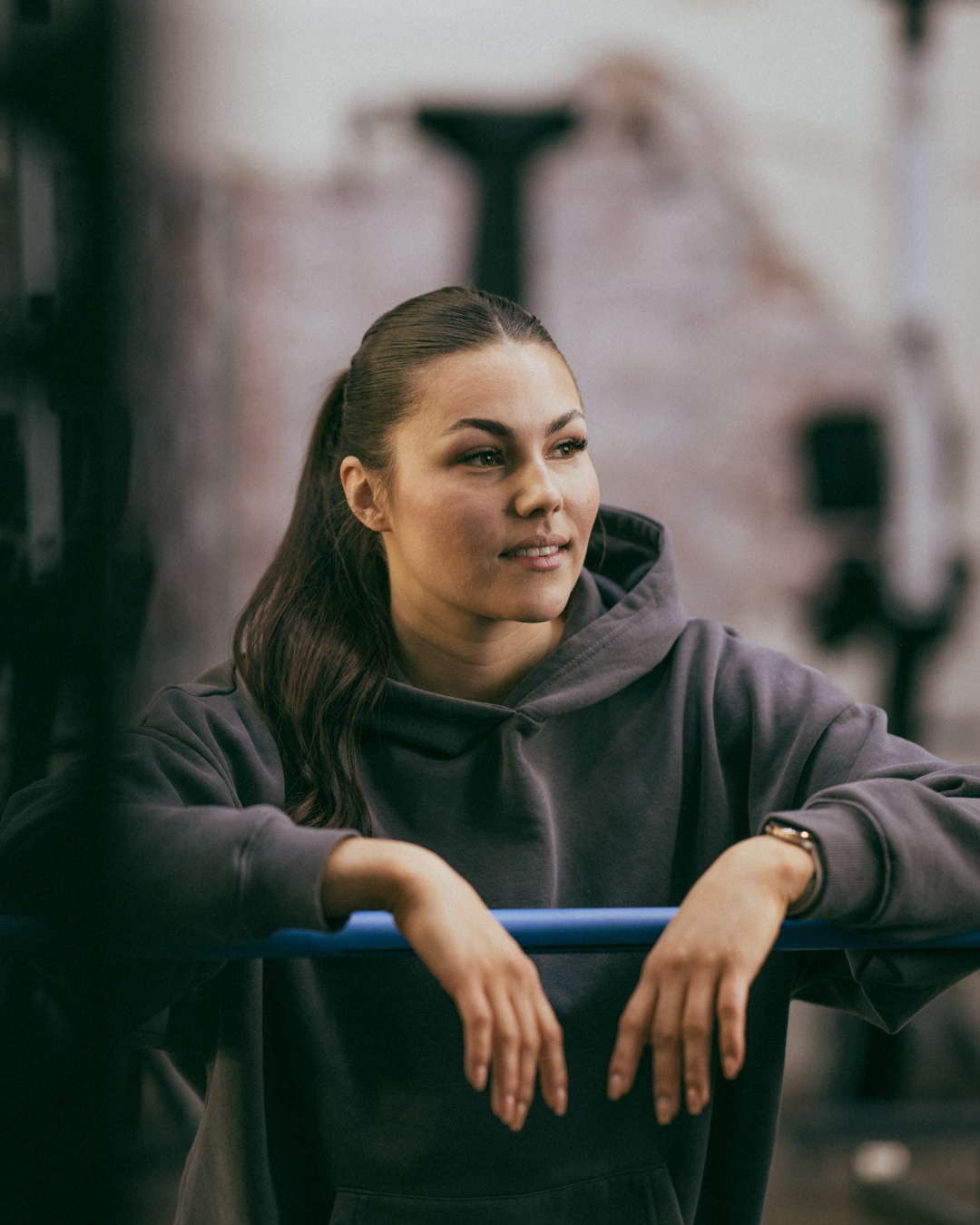 A woman in a dark hoodie rests her arms on a blue gym bar while looking away with a thoughtful expression.