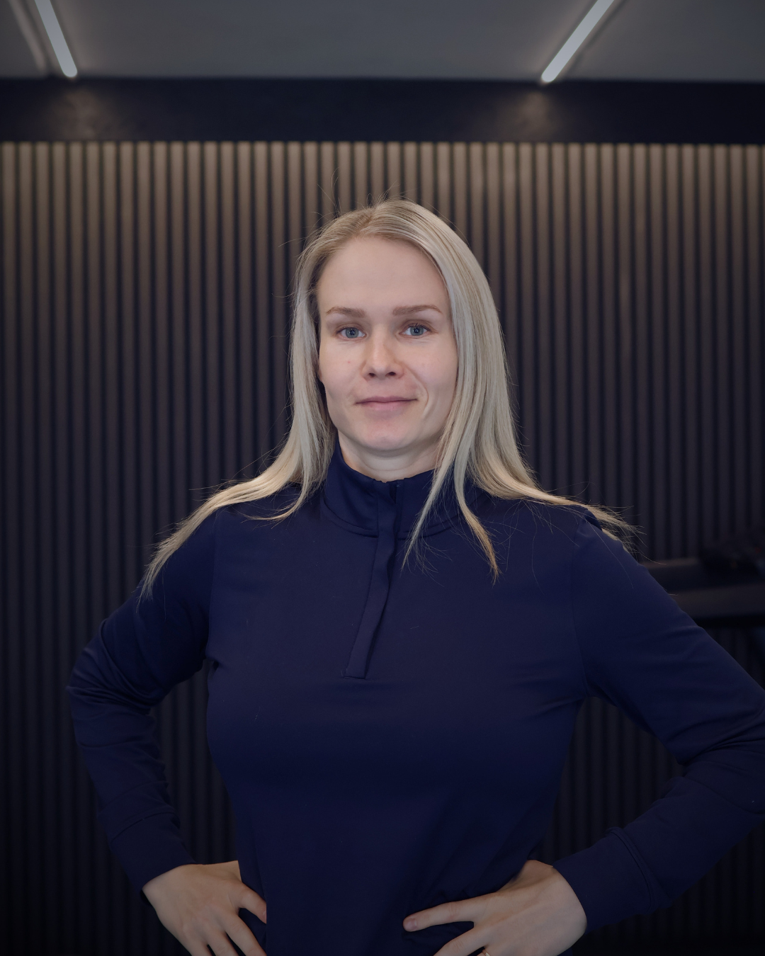 A blonde woman in a navy blue athletic top stands with her hands on her hips against a dark, slatted wall.