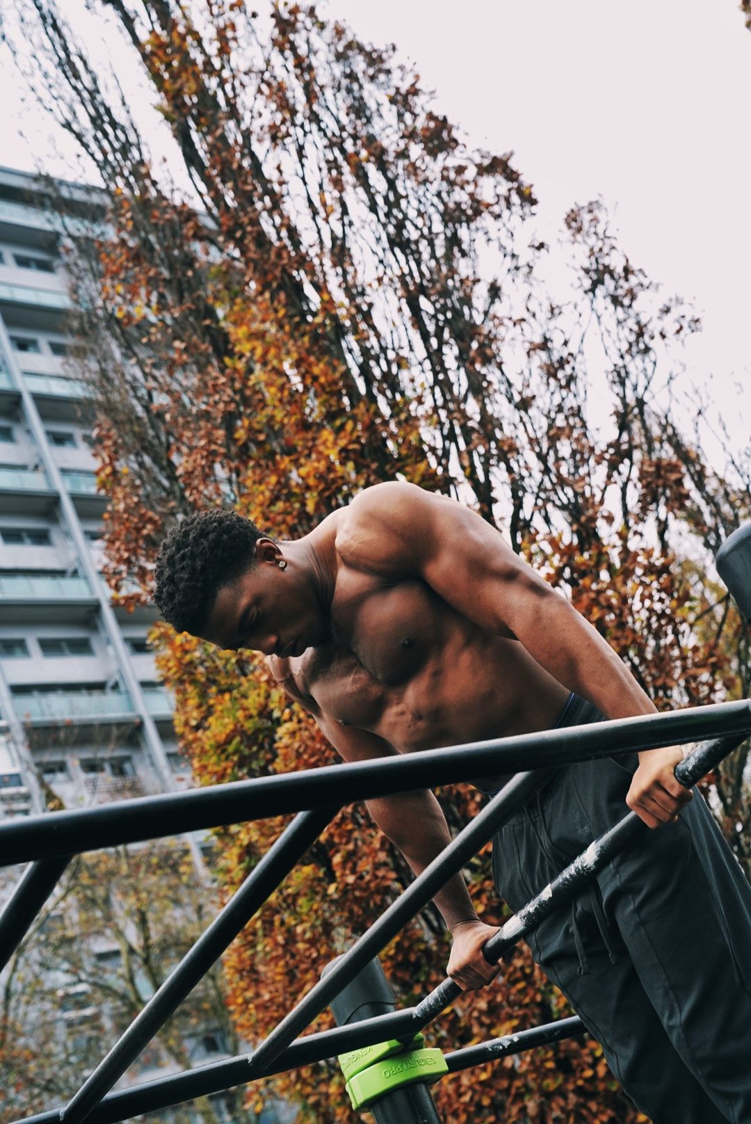 A shirtless man performs a dip exercise on parallel bars at an outdoor park with autumn trees in the background.