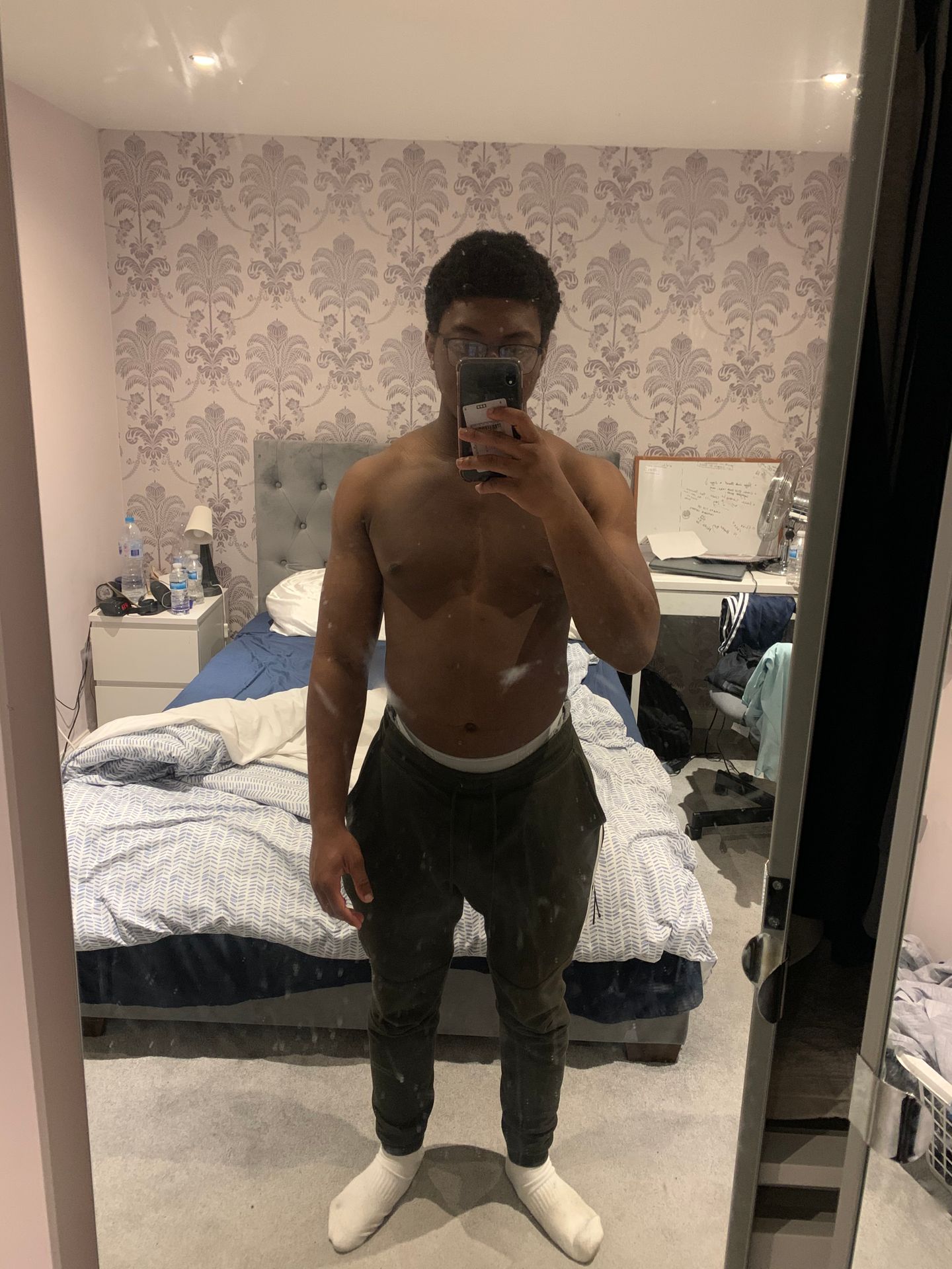A shirtless man takes a mirror selfie in his bedroom to track his fitness progress.