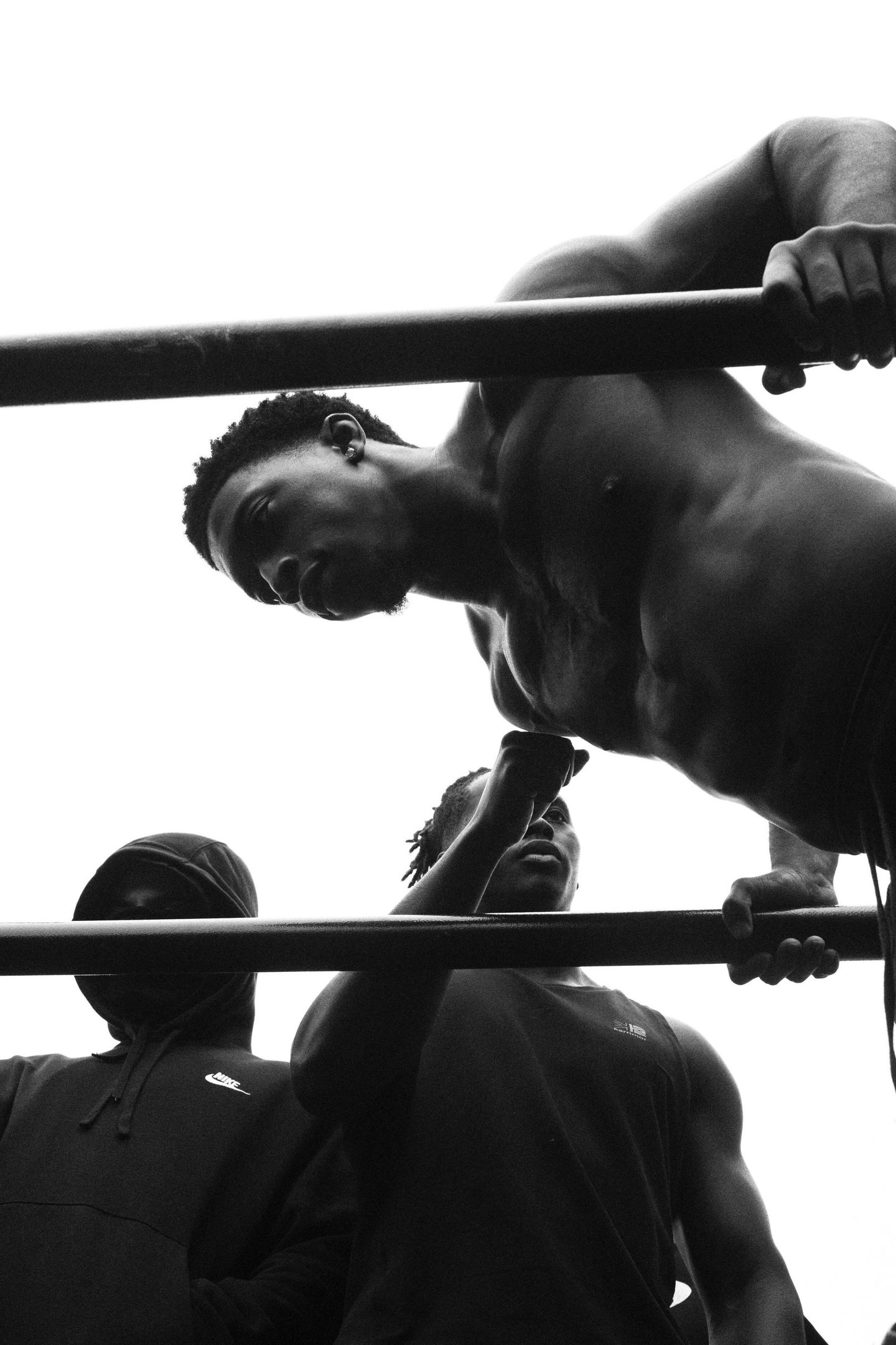 A shirtless man performs a dip on parallel bars while two other men watch from below in a low-angle black and white shot.