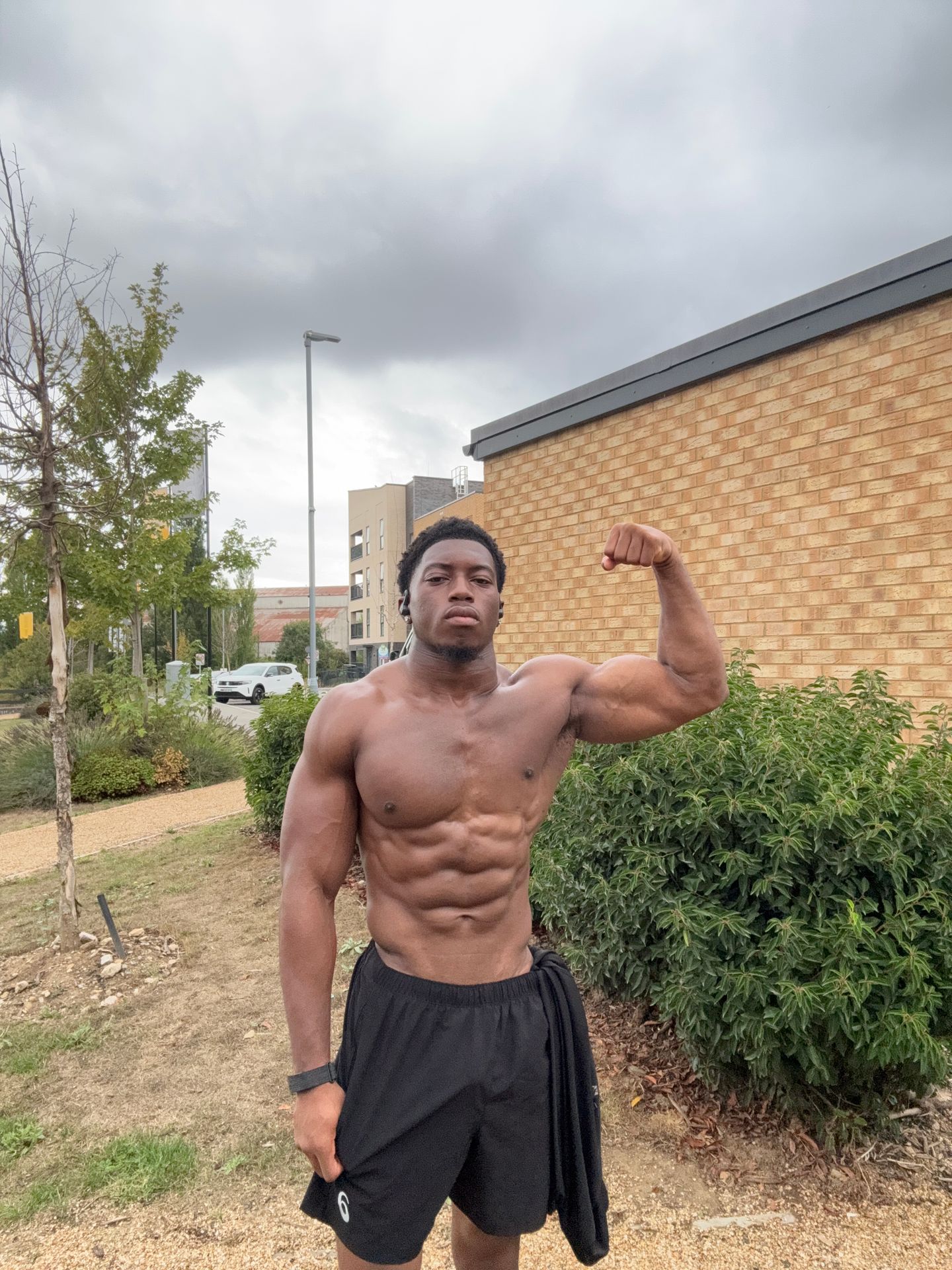 A muscular man with defined abs flexes his bicep while standing outdoors in front of a brick building.