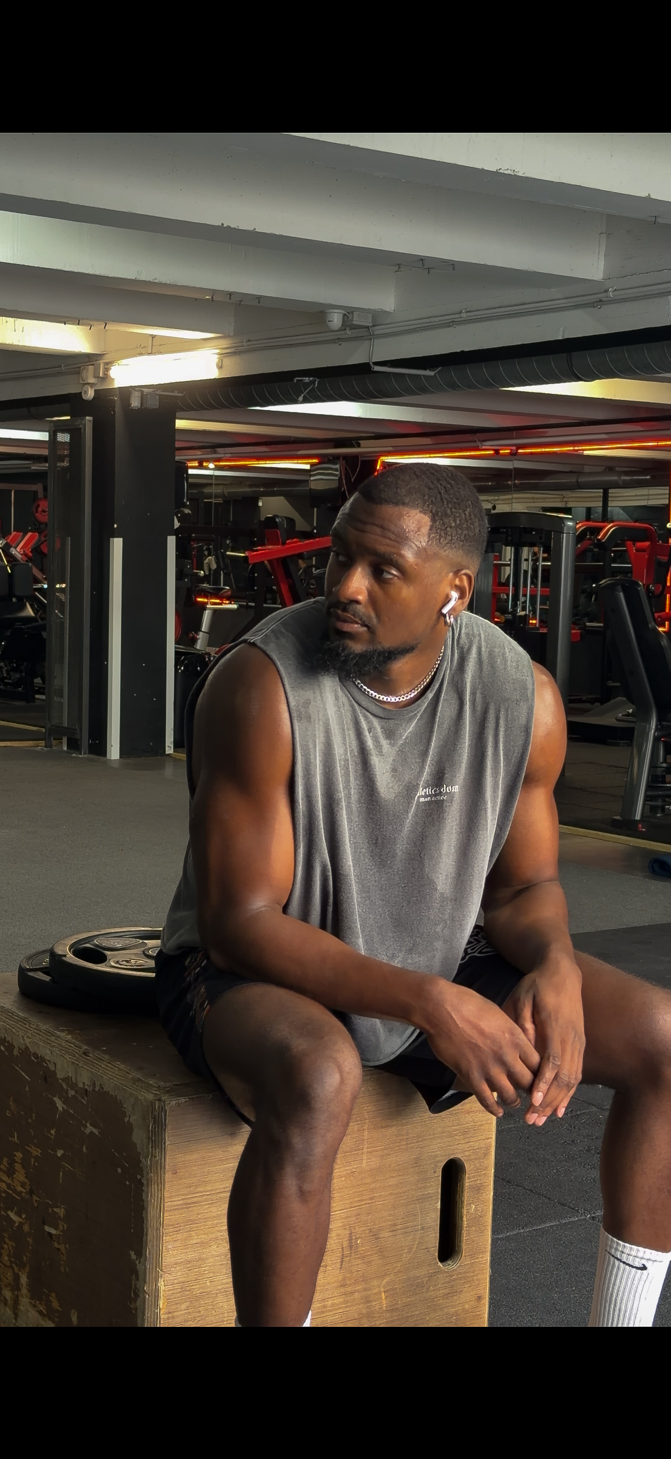 A man in a grey sleeveless shirt sits on a wooden plyometric box in a gym, looking away from the camera.