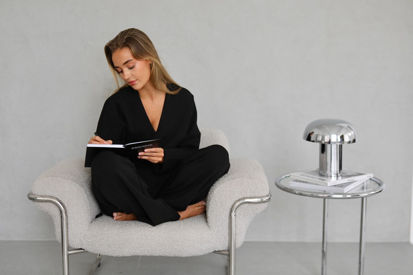 A woman in a black outfit sits cross-legged in a textured armchair, writing in a notebook.