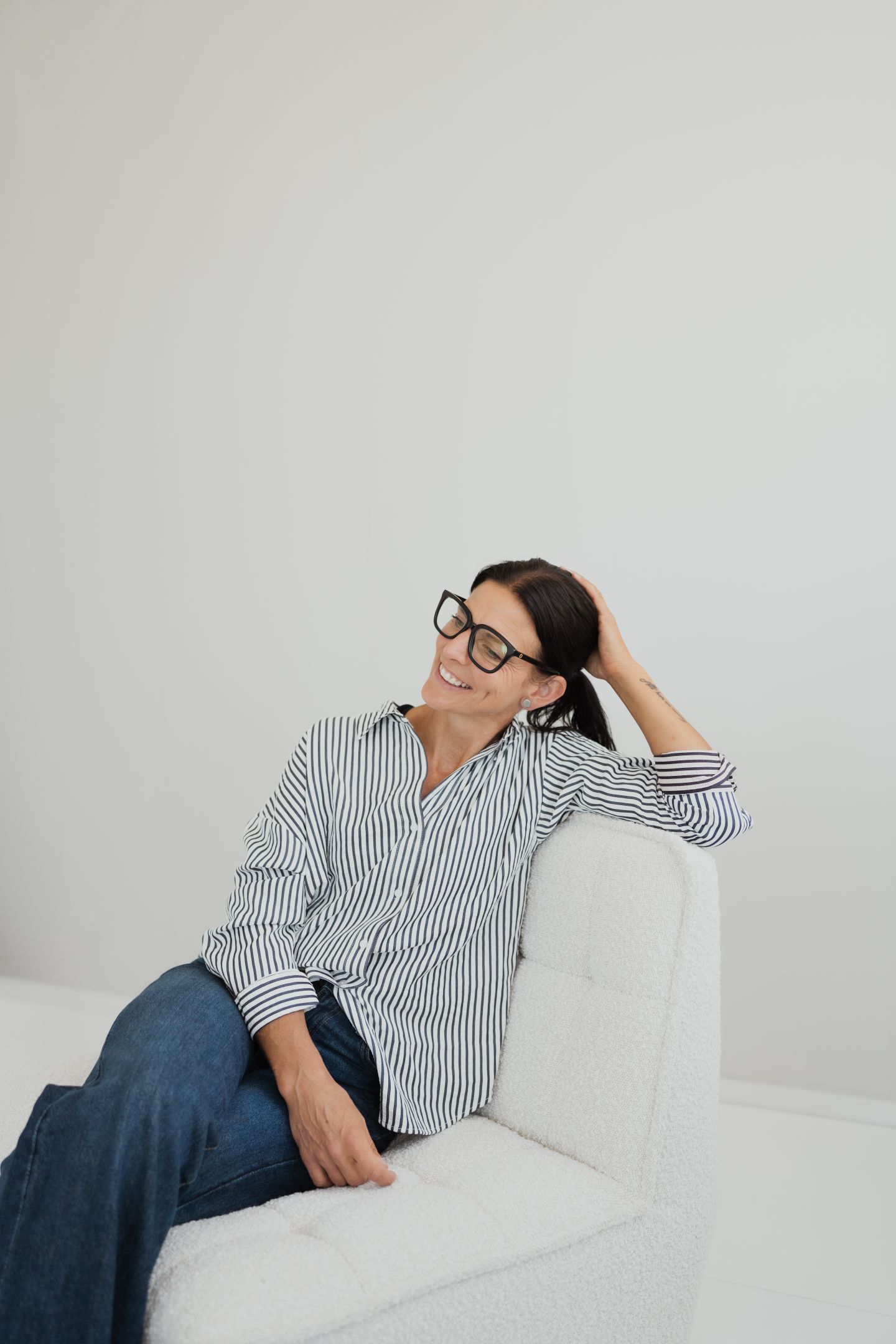A smiling woman with glasses wearing a striped shirt and jeans sits on a white chair, looking away from the camera.